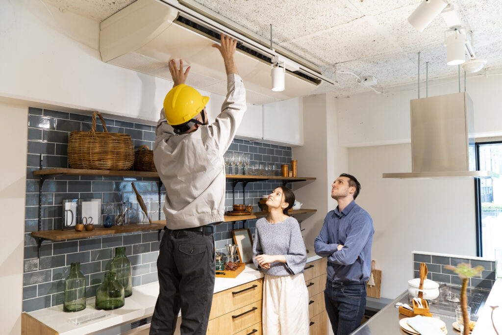 A worker in a yellow hard hat checks an air conditioning unit as two people observe in a modern kitchen.