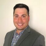 Man with short dark hair in a grey blazer and plaid shirt, smiling at the camera in front of a plain light background.