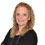 A woman with curly blonde hair, wearing a black top, smiles and faces the camera against a plain white background.