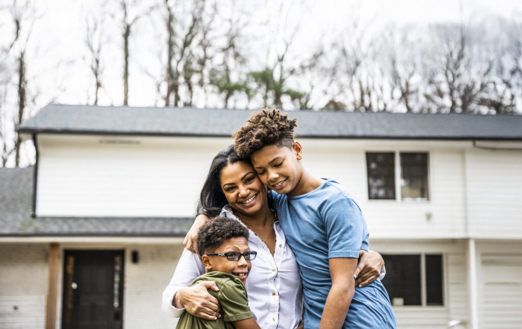 A woman stands in front of a house, smiling and hugging two boys, one in glasses and one in a blue shirt. Trees are in the background.