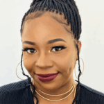 Woman with braided hair, bold makeup, gold hoop earrings, and layered necklaces, smiling in front of a light background.