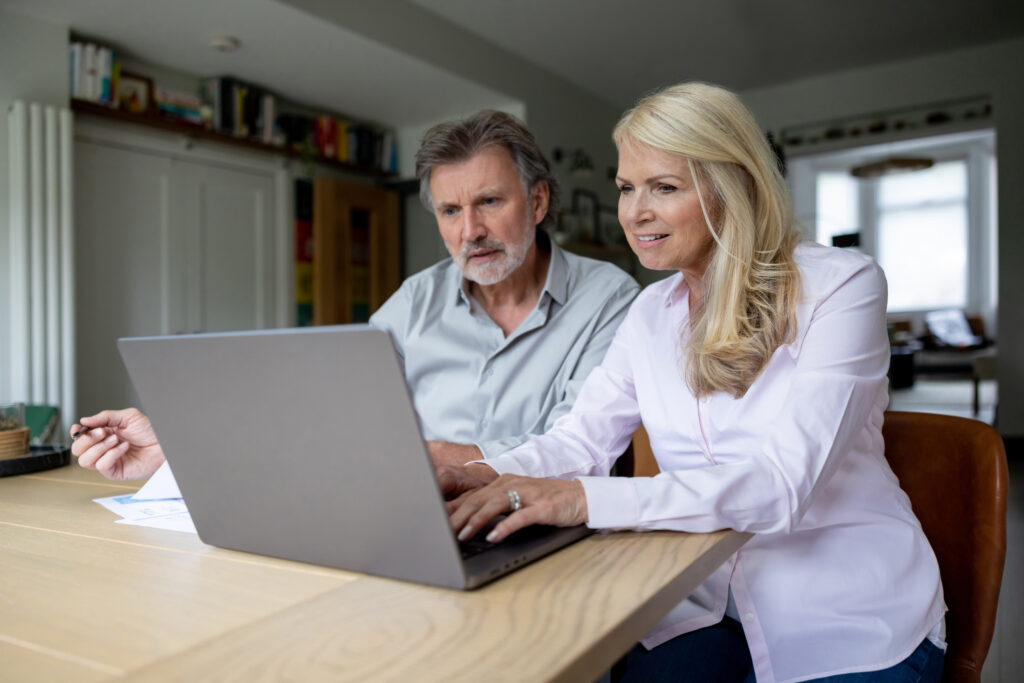 An older man and woman sit at a table, looking at a laptop screen together in a well-lit room with bookshelves in the background.
