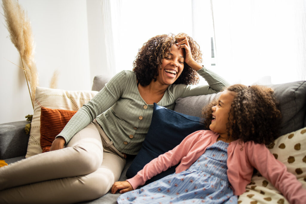 An adult and a child sit on a couch, smiling and laughing together in a brightly lit living room.