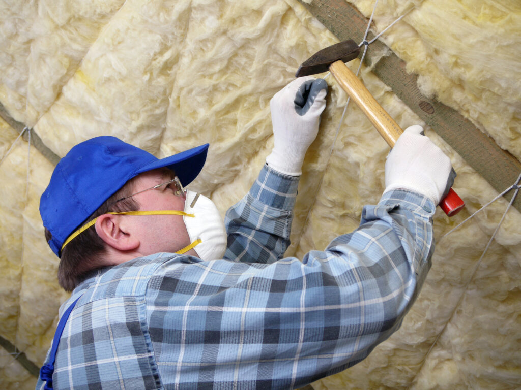 A person wearing gloves, a mask, and a cap uses a hammer to install insulation material on a ceiling.