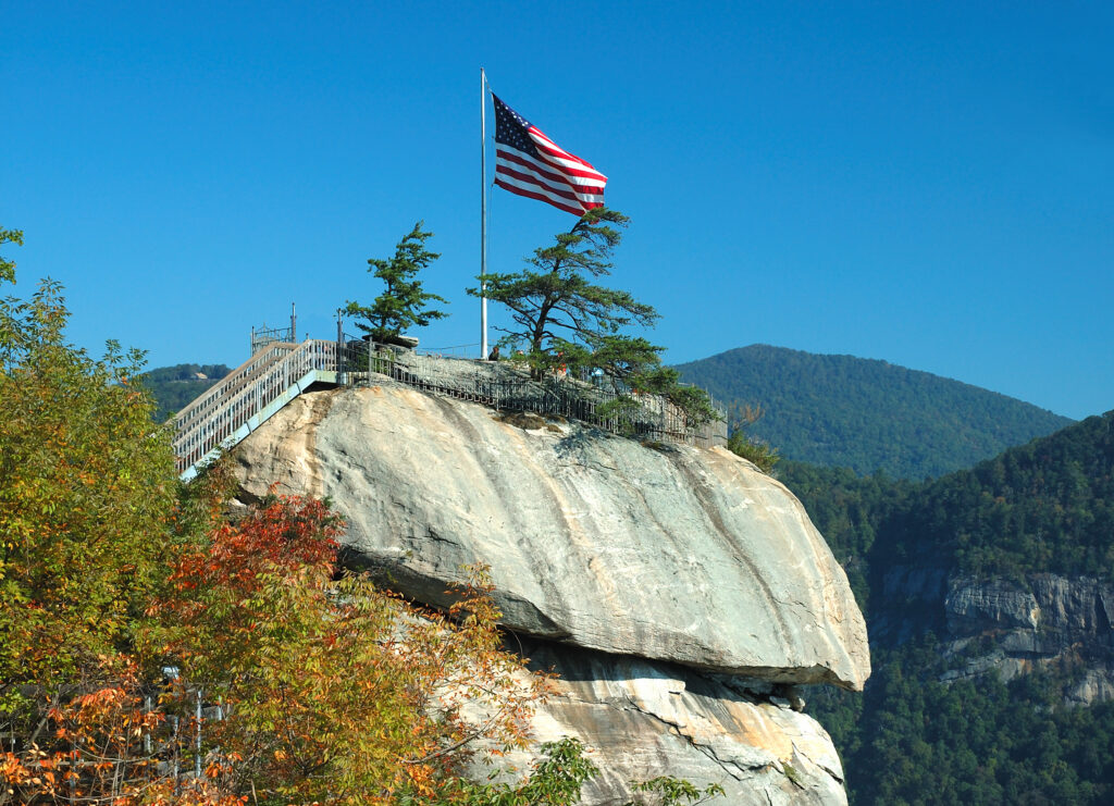 An American flag on a pole stands atop a large rock formation with a metal stairway, surrounded by trees and mountains under a clear blue sky.