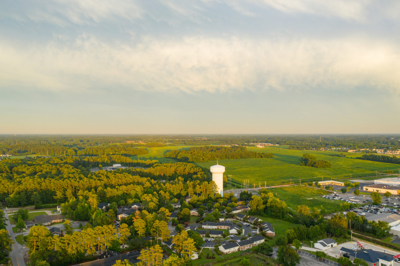 Aerial view of a suburban neighborhood with a white water tower, surrounded by trees and open green fields under a partly cloudy sky.