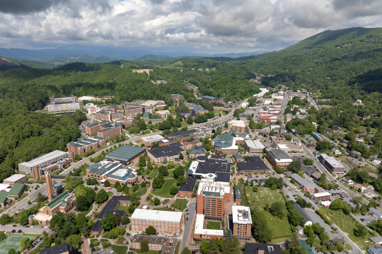Aerial view of a small city with clustered buildings, roads, green trees, and surrounding hills under a cloudy sky.