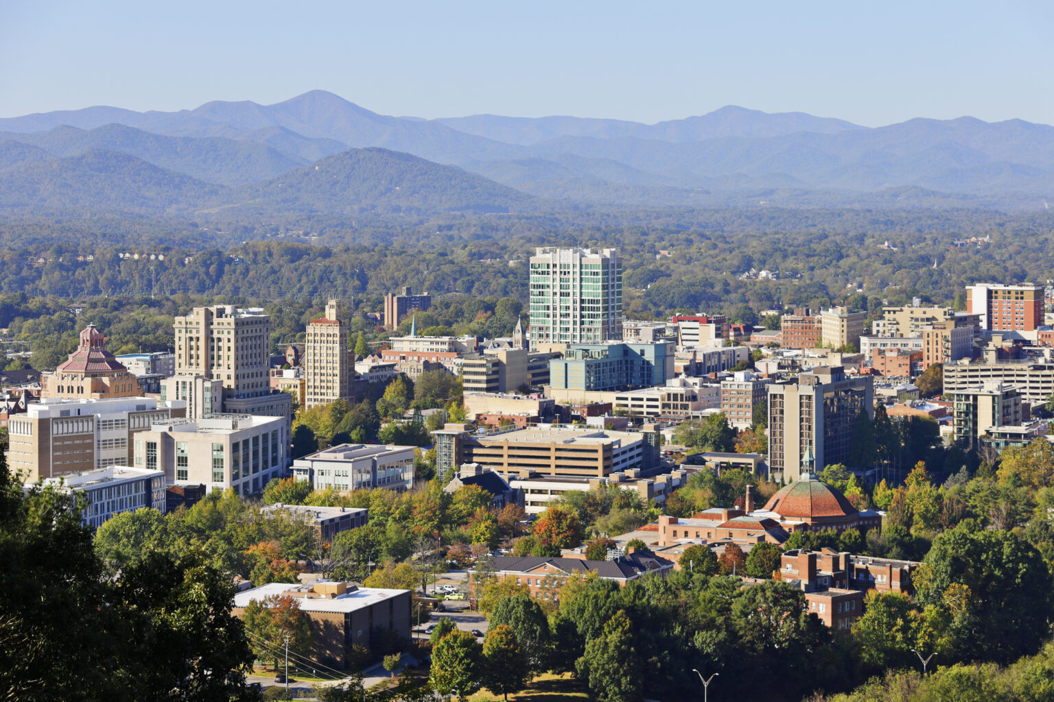 Downtown cityscape with mid-rise buildings, surrounded by trees and mountains in the background under a clear blue sky.