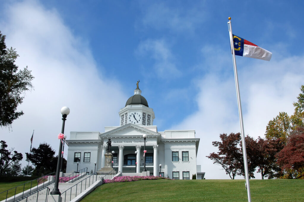 A white courthouse with columns and a clock tower sits on a hill; a flagpole with a North Carolina flag stands in the foreground.