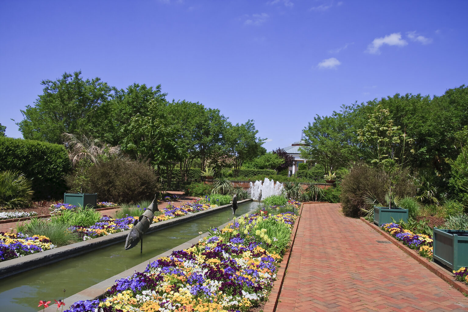 A brick walkway runs beside a flower-filled garden bed with a water feature and bird sculptures, surrounded by green trees under a blue sky.