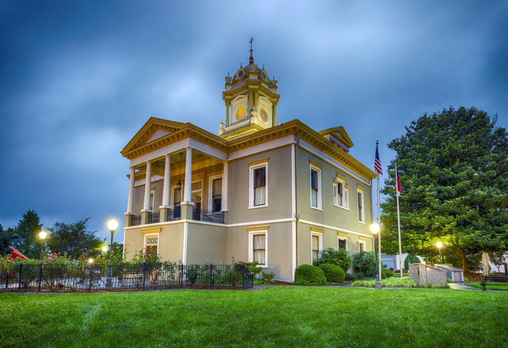 Historic courthouse building with columns and a clock tower, surrounded by grass, trees, and lampposts, taken at dusk.