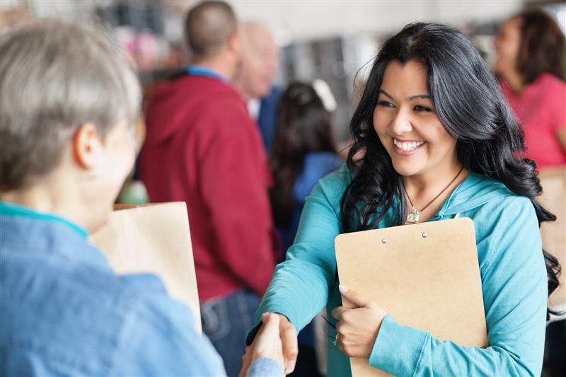 Woman holding a clipboard smiles and shakes hands with another person in a busy indoor setting with several people in the background.