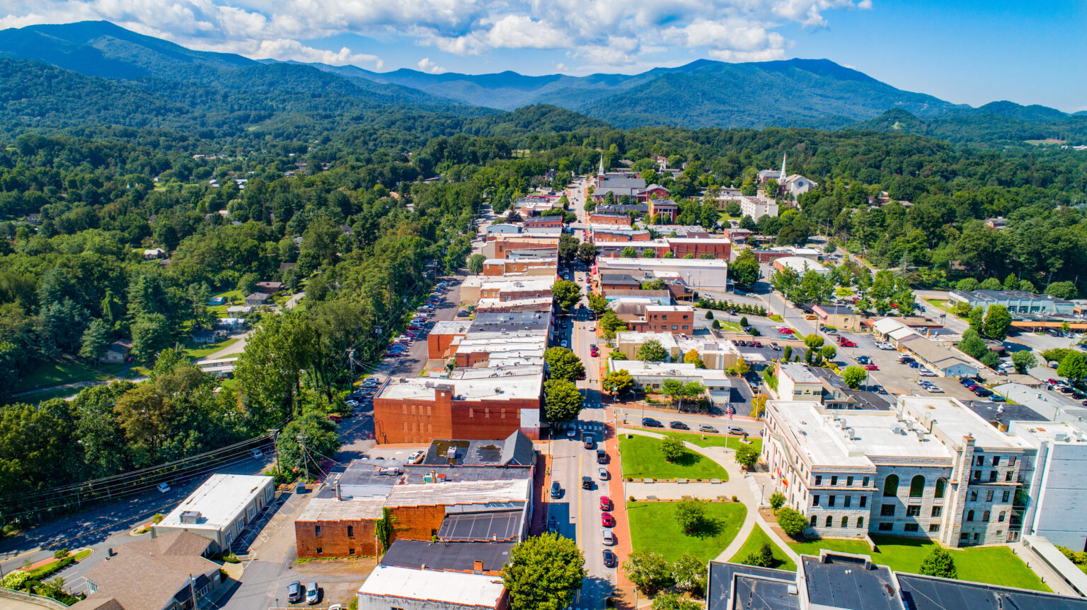 Aerial view of a small town with brick buildings, green spaces, and mountains in the background under a partly cloudy sky.