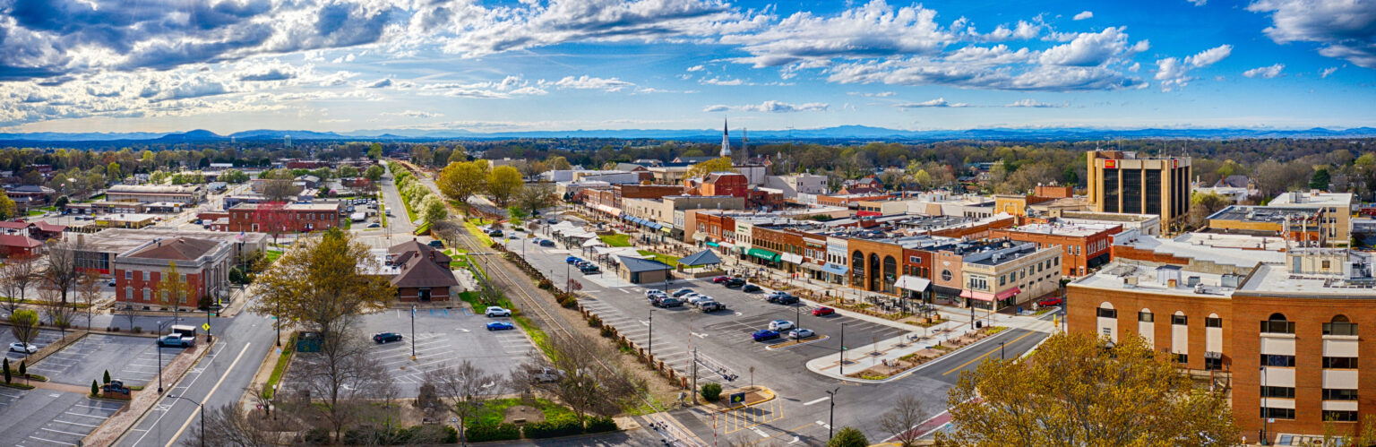 Aerial view of a small town’s main street with shops, parking lots, trees, and distant mountains under a partly cloudy sky.