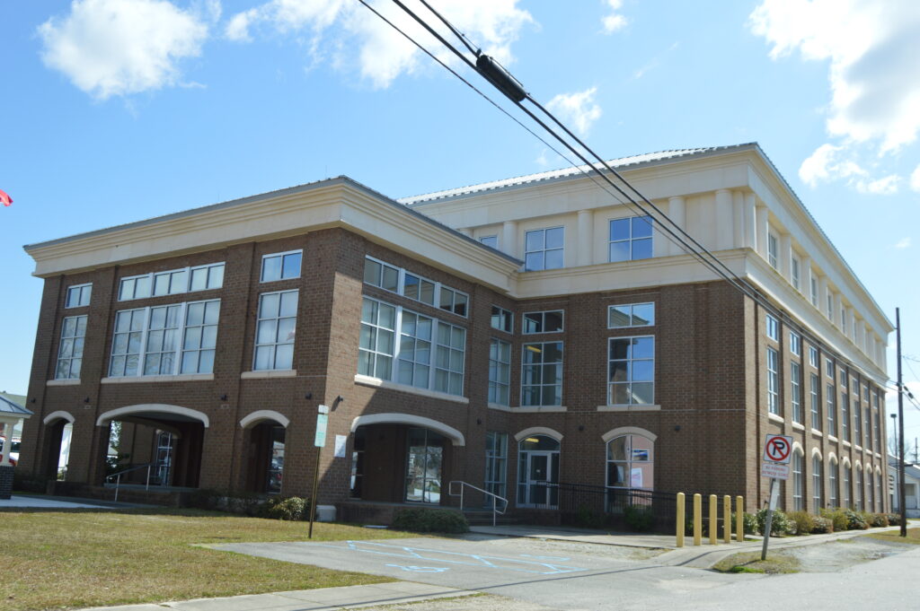 Three-story brick building with large windows, arched entrances, and a sloped roof, situated beside a street with a no parking sign.