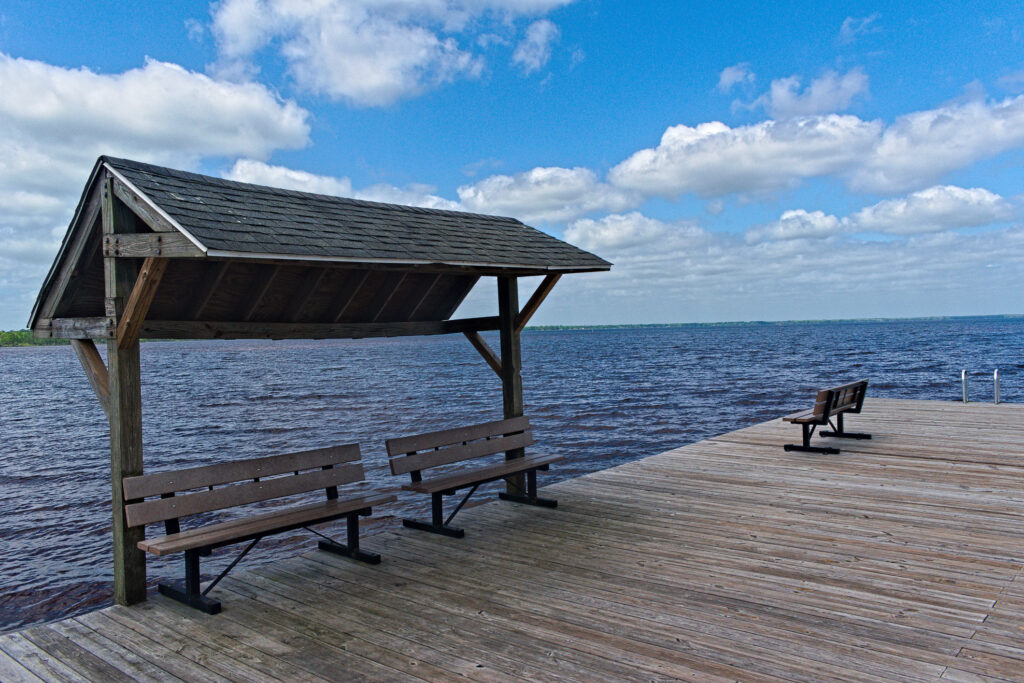 A wooden shelter with benches sits on a lakeside dock under a partly cloudy sky; another bench is on the open dock.