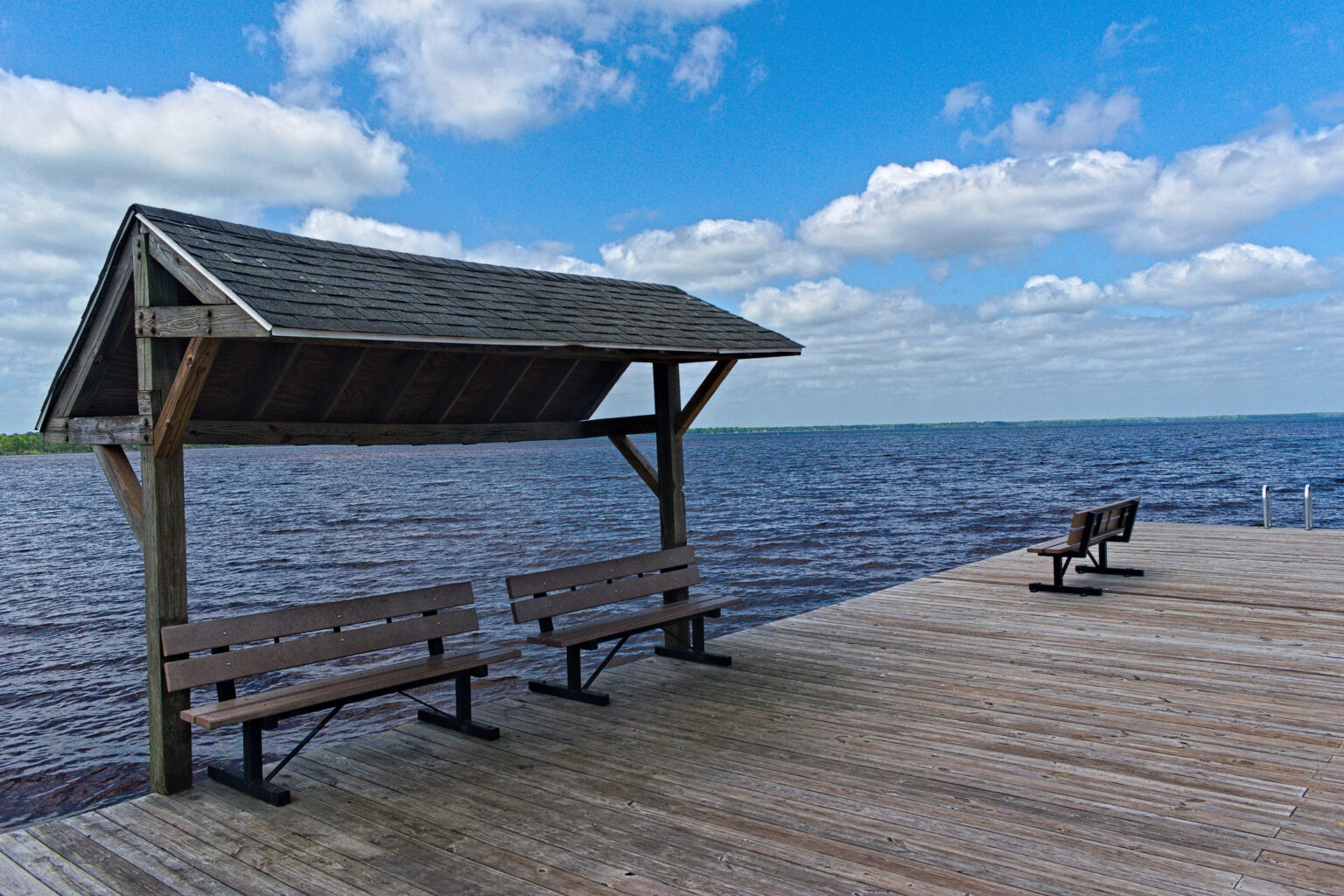 A wooden shelter with benches sits on a lakeside dock under a partly cloudy sky; another bench is on the open dock.
