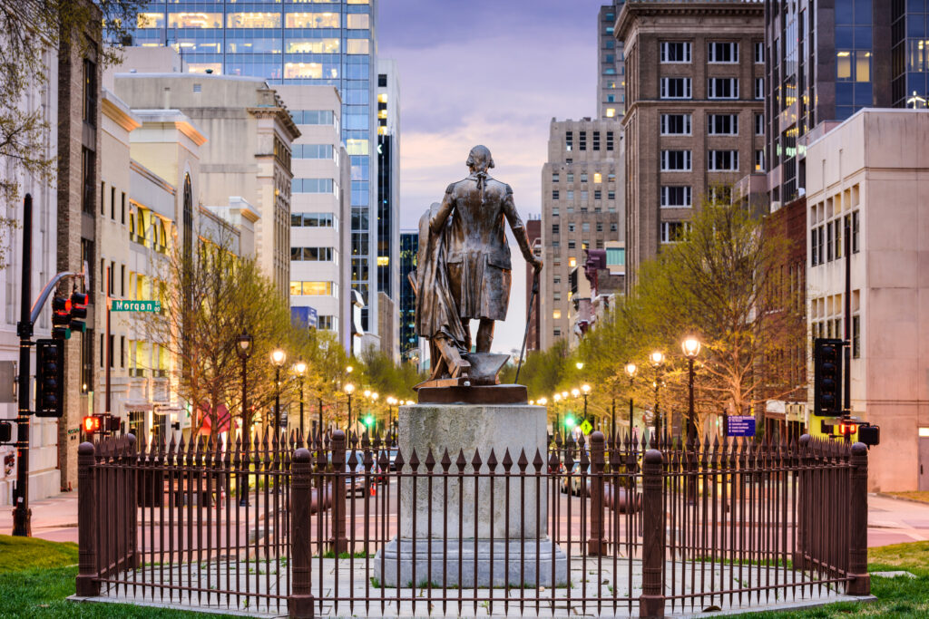A bronze statue of a man stands on a pedestal behind a fence, surrounded by buildings and trees on a city street at dusk.