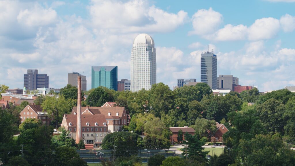 City skyline with tall modern buildings in the background and red-roofed brick buildings and trees in the foreground under a partly cloudy sky.