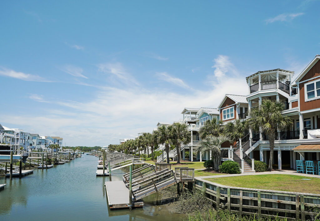 Waterfront houses with docks line a calm canal under a blue sky with scattered clouds and palm trees along the shore.