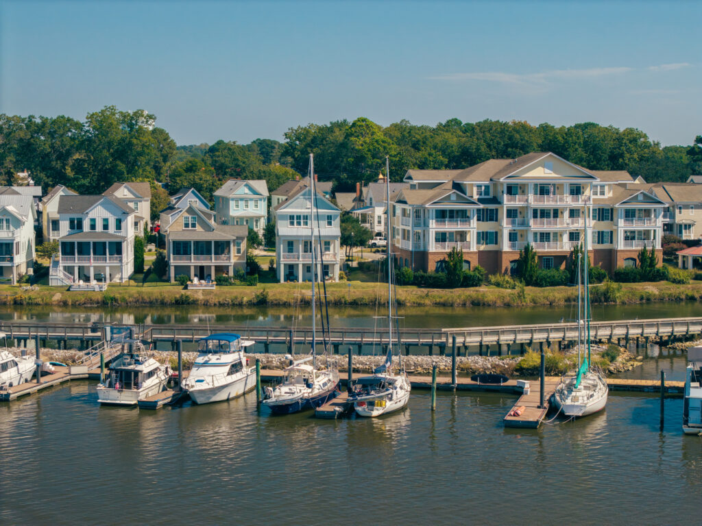 Several boats and yachts are docked at a marina with waterfront houses and apartment buildings in the background.