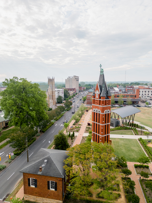 Aerial view of a city street with a red brick tower, a church, trees, and buildings under a cloudy sky.