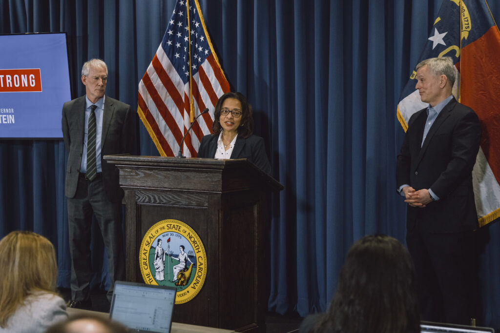 Three people stand at a podium with North Carolina and US flags; one woman speaks while two men stand on either side. Audience members face them.