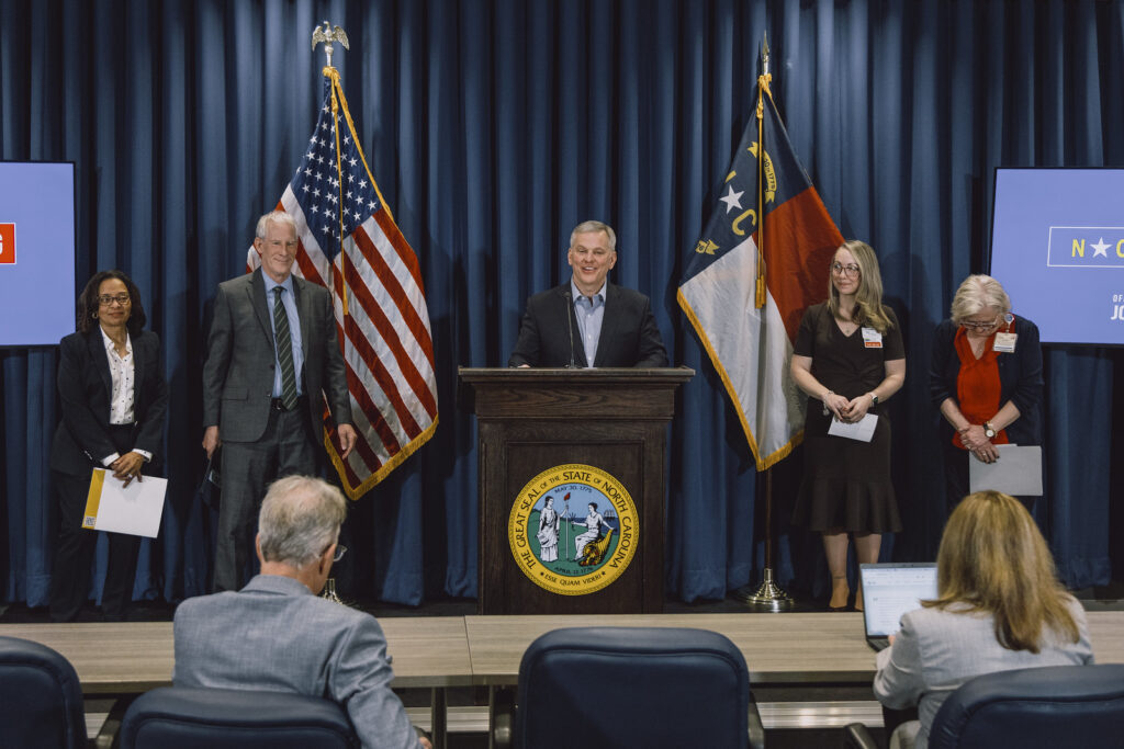 Five people stand on a stage behind a podium with the North Carolina state seal, with two flags and blue curtains in the background.