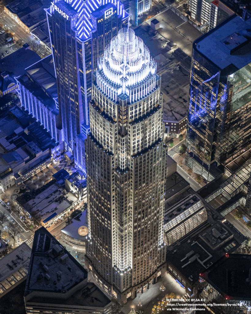 Aerial view of a tall, illuminated skyscraper with a crown-like top in a city at night, surrounded by other lit buildings.