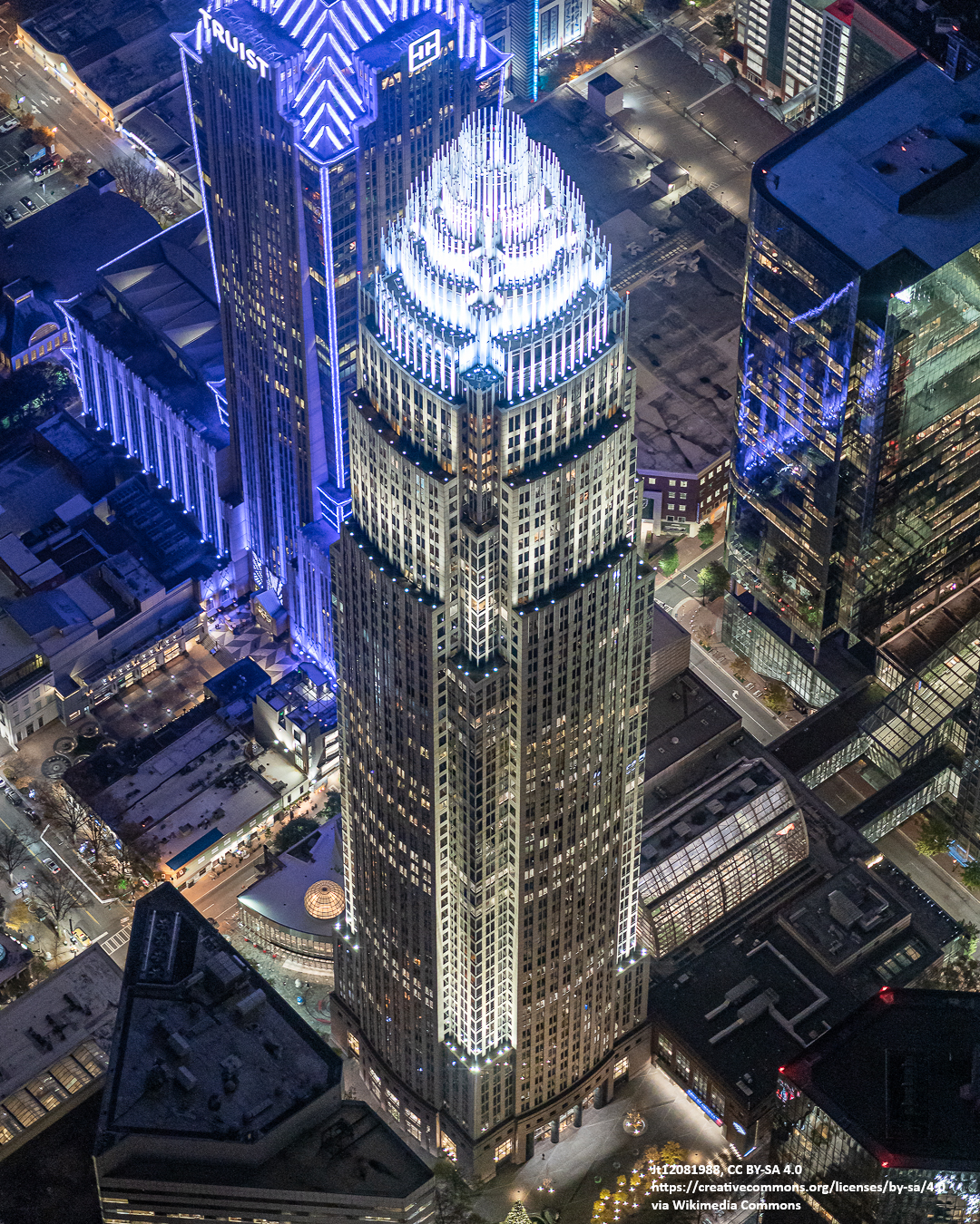 Aerial view of a tall, illuminated skyscraper with a crown-like top in a city at night, surrounded by other lit buildings.