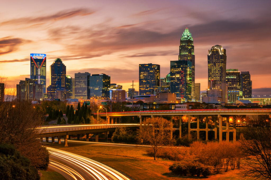 Charlotte, North Carolina skyline at sunset with light trails on a freeway and buildings illuminated against a colorful sky.